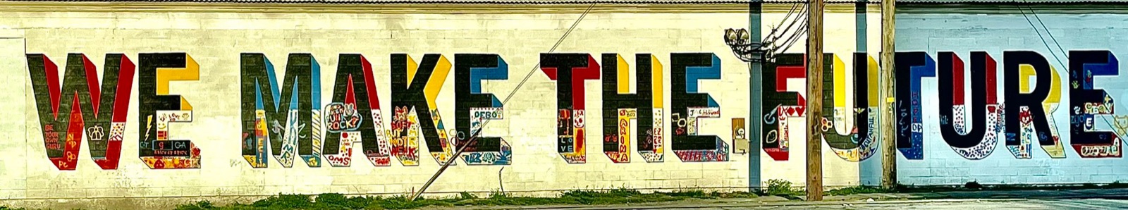 A long cinder-block wall painted with the words WE MAKE THE FUTURE in tall multicolored block letters, each letter filled with small hand-drawn doodles, with a wooden utility pole standing in front